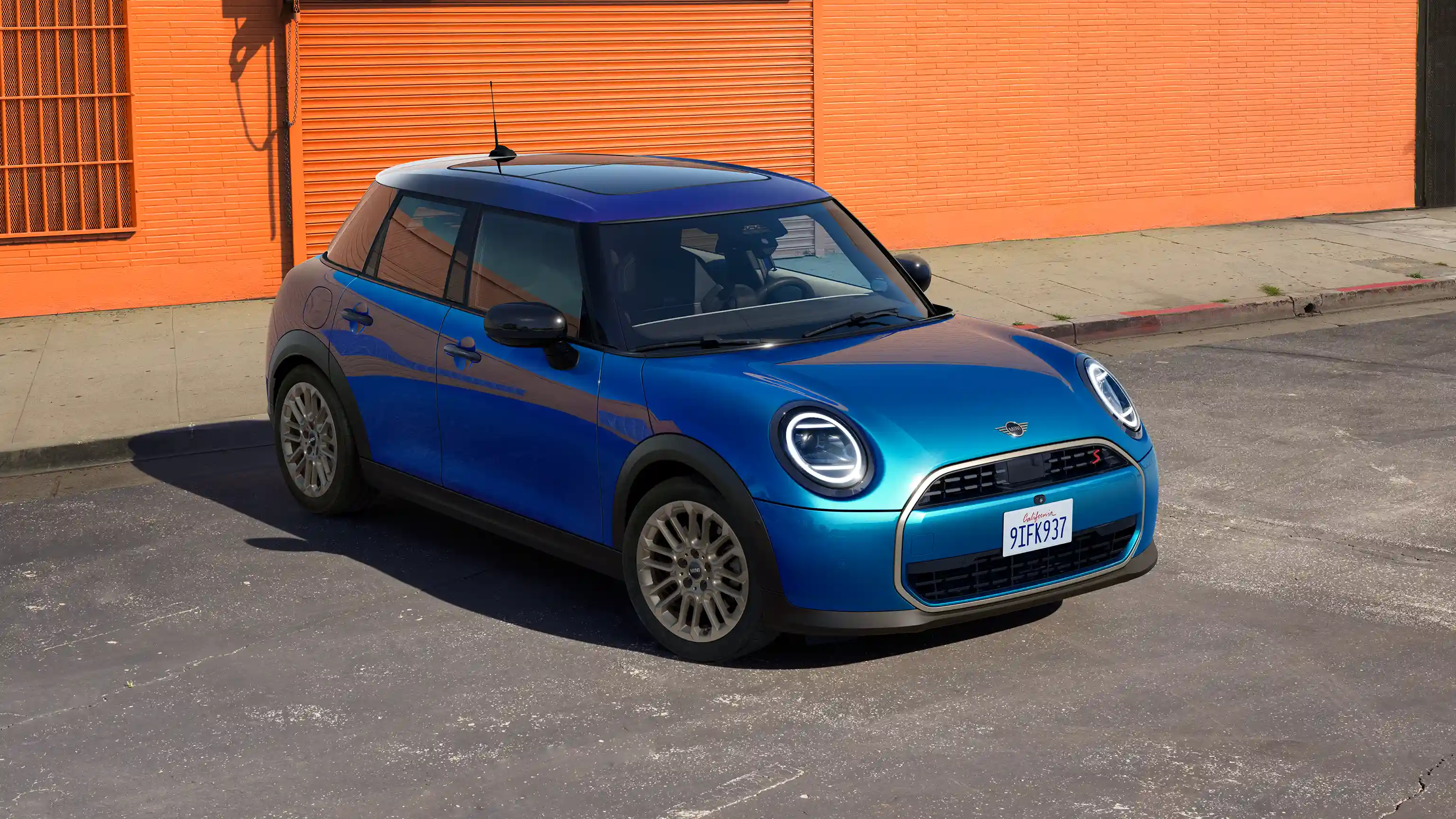 Frontansicht eines parkenden MINI Cooper 5-Türer in Icy Sunshine Blue mit Dach mit Multitone-Farbverlauf in Blau auf einer Straße mit einer orangefarbenen Wand im Hintergrund