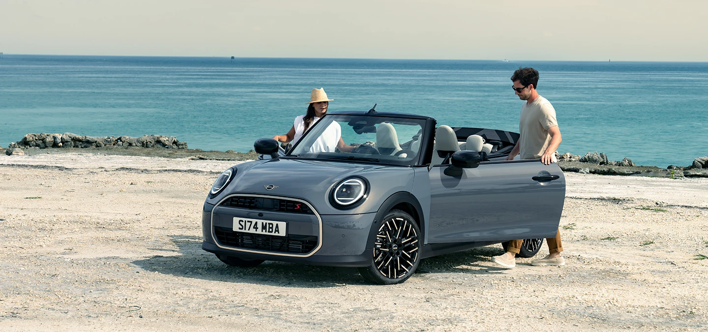 Vue avant gauche de la MINI Cooper Cabrio Cooper Grey au bord d’une plage avec deux personnes à côté.