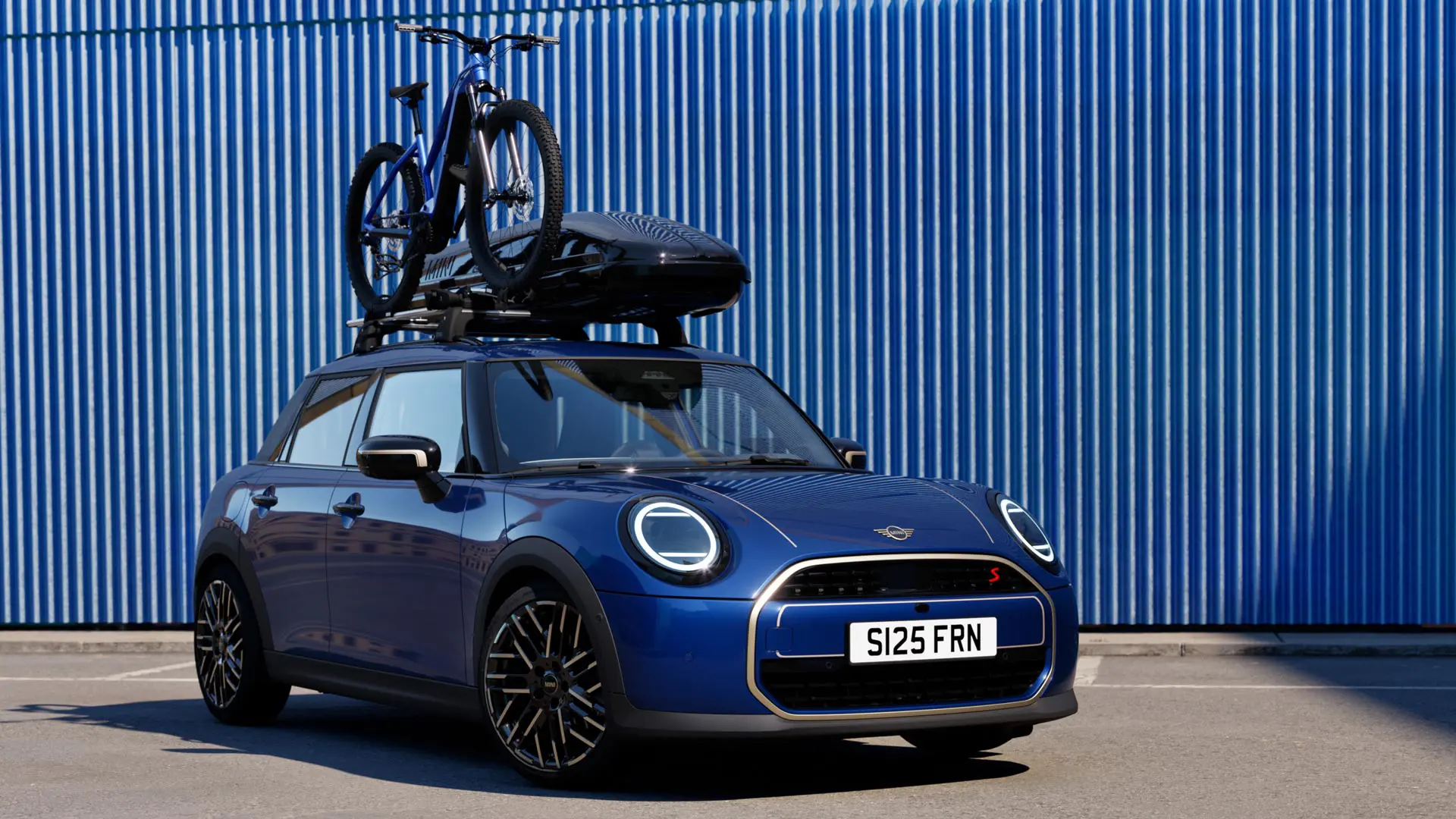 Vorderansicht des MINI Aceman in Indigo Sunset Blue mit MINI Fahrraddachträger mit Mountainbike und MINI Dachbox 320.
